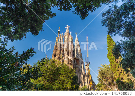 Sagrada Familia basilica rising above green trees in Barcelona under dramatic blue summer sky Sagrada Familia basilica rising above green trees in Barcelona under dramatic blue summer sky 141768033