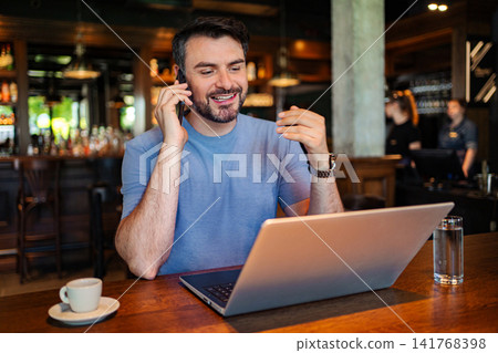 Man working remotely on laptop making phone call in cafe Man working remotely on laptop making phone call in cafe 141768398