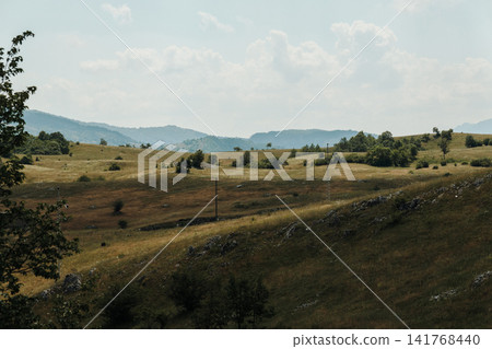Rolling grassy hills and distant mountain ridges create a peaceful rural landscape in Montenegro. The scenic countryside is bathed in soft summer light beneath dramatic clouds Rolling grassy hills and distant mountain ridges create a peaceful rural landscape in Montenegro. The scenic countryside is bathed in soft summer light beneath dramatic clouds 141768440
