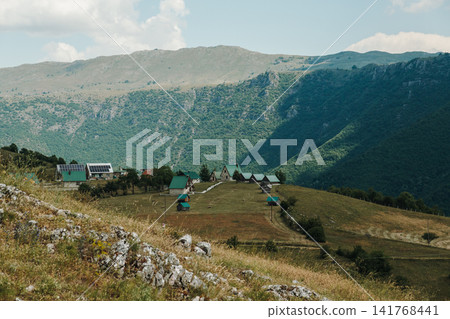 A small mountain village sits on a grassy hillside surrounded by dramatic peaks in Montenegro. Traditional rural buildings overlook a peaceful alpine landscape during summer A small mountain village sits on a grassy hillside surrounded by dramatic peaks in Montenegro. Traditional rural buildings overlook a peaceful alpine landscape during summer 141768441