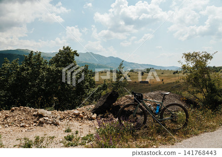 A loaded gravel bicycle stands beside a mountain viewpoint overlooking the rolling countryside of Montenegro. The peaceful landscape reflects the spirit of bikepacking adventure A loaded gravel bicycle stands beside a mountain viewpoint overlooking the rolling countryside of Montenegro. The peaceful landscape reflects the spirit of bikepacking adventure 141768443