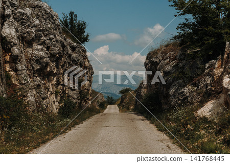 A narrow mountain road cuts through a rocky pass surrounded by limestone cliffs in Montenegro. The scenic landscape leads toward distant mountain peaks under a summer sky 141768445
