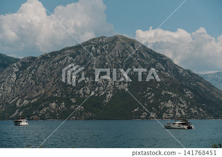 Modern catamaran anchored on the calm waters of the Bay of Kotor in Montenegro. Rocky mountains and dramatic clouds create a scenic Adriatic backdrop Modern catamaran anchored on the calm waters of the Bay of Kotor in Montenegro. Rocky mountains and dramatic clouds create a scenic Adriatic backdrop 141768451