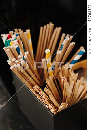 Paper drinking straws and wooden stirrers neatly organized on a coffee shop counter. Eco-friendly accessories for serving beverages Paper drinking straws and wooden stirrers neatly organized on a coffee shop counter. Eco-friendly accessories for serving beverages 141768502