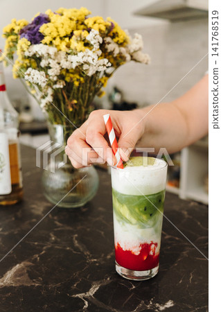 Hand placing a striped paper straw into a freshly prepared layered strawberry matcha latte. Refreshing iced cafe beverage ready to serve 141768519
