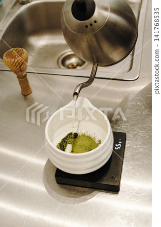 Hot water being poured over sifted matcha powder in a ceramic bowl on a digital scale. Traditional Japanese tea preparation in progress Hot water being poured over sifted matcha powder in a ceramic bowl on a digital scale. Traditional Japanese tea preparation in progress 141768535