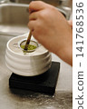 Bamboo whisk gently presses matcha powder through a fine mesh strainer into a ceramic bowl. Traditional tea preparation captured in close-up 141768536