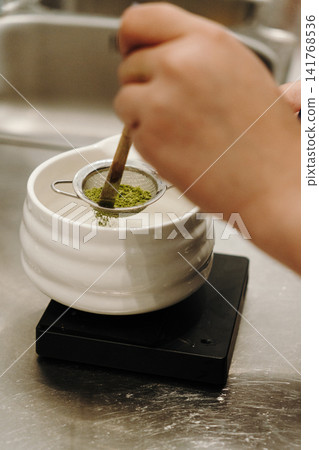 Bamboo whisk gently presses matcha powder through a fine mesh strainer into a ceramic bowl. Traditional tea preparation captured in close-up 141768536