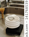 Fine mesh tea strainer resting on a ceramic bowl placed on a digital kitchen scale. Coffee and tea preparation tools arranged on a stainless steel countertop 141768539