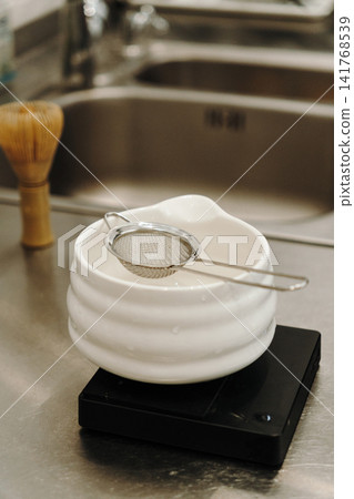 Fine mesh tea strainer resting on a ceramic bowl placed on a digital kitchen scale. Coffee and tea preparation tools arranged on a stainless steel countertop Fine mesh tea strainer resting on a ceramic bowl placed on a digital kitchen scale. Coffee and tea preparation tools arranged on a stainless steel countertop 141768539