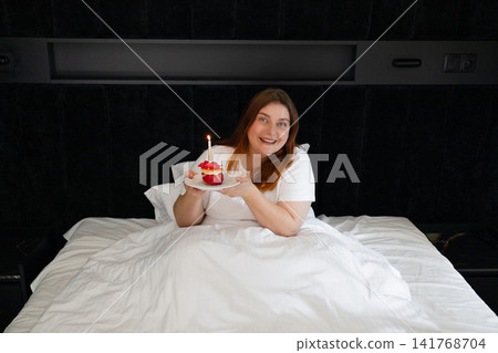 Happy young woman celebrating birthday in bed with dessert and candle Happy young woman celebrating birthday in bed with dessert and candle 141768704