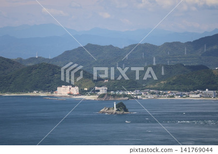 A landscape featuring the blue ocean, white waves, a small island with a lighthouse, the coastline, a townscape, a range of mountains, and a cloudy sky. A landscape featuring the blue ocean, white waves, a small island with a lighthouse, the coastline, a townscape, a range of mountains, and a cloudy sky. 141769044