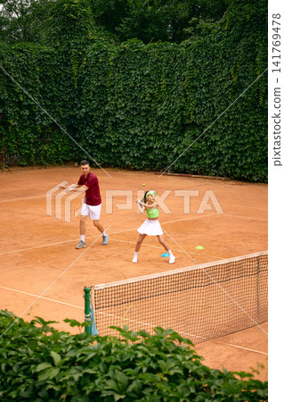 Young girl practicing tennis strokes with male coach during outdoor training session Young girl practicing tennis strokes with male coach during outdoor training session 141769478