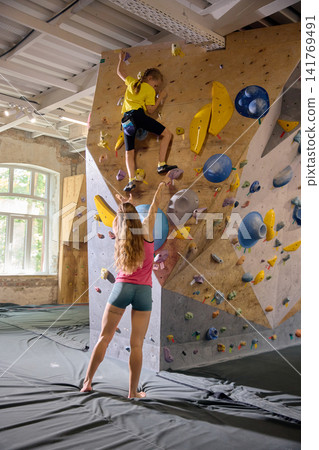 Young girl climbing high bouldering wall as female coach encourages from safety mat Young girl climbing high bouldering wall as female coach encourages from safety mat 141769491