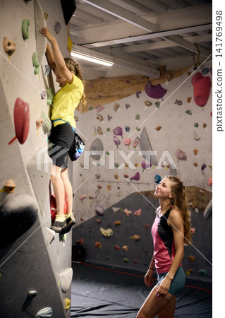 Young girl independently climbing indoor bouldering route while female coach observes Young girl independently climbing indoor bouldering route while female coach observes 141769498