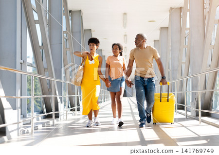 Black parents and daughter walk hand in hand through airport with luggage Black parents and daughter walk hand in hand through airport with luggage 141769749