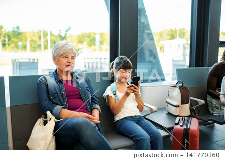 Grandmother and granddaughter waiting for travel in airport terminal Grandmother and granddaughter waiting for travel in airport terminal 141770160