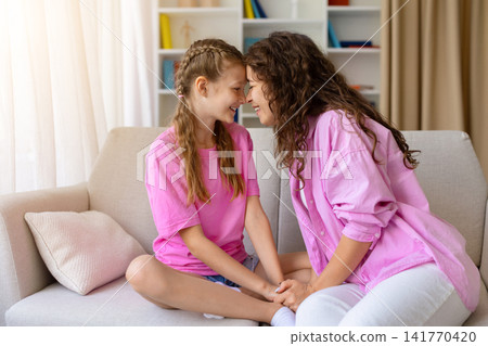 Happy mother and daughter sitting together on sofa, touching foreheads Happy mother and daughter sitting together on sofa, touching foreheads 141770420