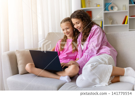 Smiling mother and daughter sitting together on sofa using laptop at home Smiling mother and daughter sitting together on sofa using laptop at home 141770554