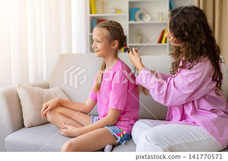 Caring mother braiding her daughters hair while they sit together on sofa Caring mother braiding her daughters hair while they sit together on sofa 141770571