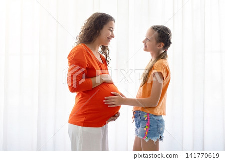 Pregnant mother and young daughter standing by bright window, smiling and bonding Pregnant mother and young daughter standing by bright window, smiling and bonding 141770619