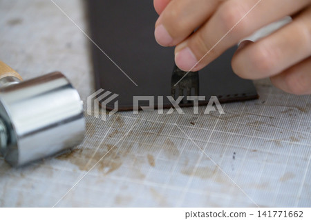 A leather craftsman's hands making sewing holes in leather with a four-pronged diamond awl. A leather craftsman's hands making sewing holes in leather with a four-pronged diamond awl. 141771662