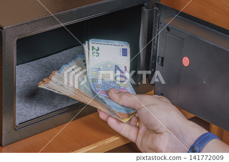 Person hand taking euro cash banknotes out of open safe box placed on wooden shelf inside hotel room cabinet for financial security asset storage concept Person hand taking euro cash banknotes out of open safe box placed on wooden shelf inside hotel room cabinet for financial security asset storage concept 141772009