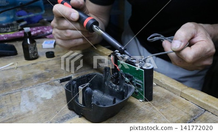 A mechanic repairs a battery pack in a workshop. He holds a soldering iron, carefully soldering contacts to the electronic circuit board with wires, and a gray cable, working at a workbench. A mechanic repairs a battery pack in a workshop. He holds a soldering iron, carefully soldering contacts to the electronic circuit board with wires, and a gray cable, working at a workbench. 141772020