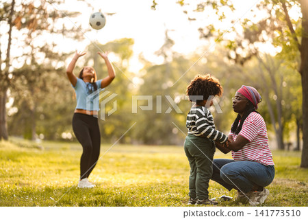 Mother and daugther playing football with child watching in park Mother and daugther playing football with child watching in park 141773510