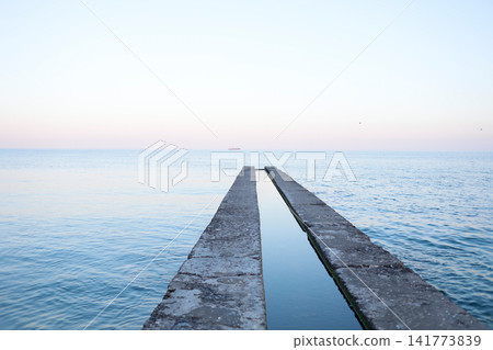 Minimalist view of a long concrete pier extending into calm blue sea beneath a clear sky, emphasizing symmetry, tranquil water, coastal scenery and peaceful marine horizon vistas Minimalist view of a long concrete pier extending into calm blue sea beneath a clear sky, emphasizing symmetry, tranquil water, coastal scenery and peaceful marine horizon vistas 141773839