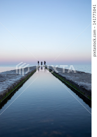 Symmetrical view of a long concrete pier stretching into calm sea at sunset, with three people at the end, capturing peaceful horizon, reflections and serene coastal landscape sea 141773841