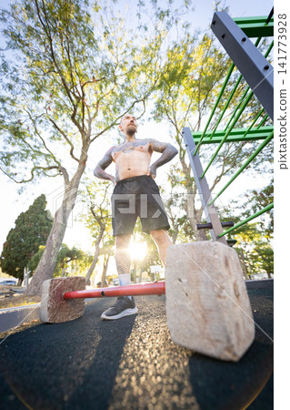 Man stands by weights in outdoor park during bright day Man stands by weights in outdoor park during bright day 141773928