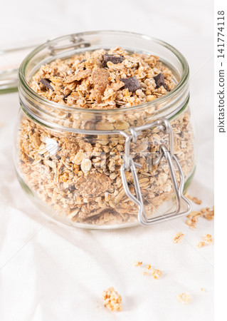 A glass jar filled with crunchy granola and chocolate chunks rests on a white cloth, a perfect healthy and wholesome breakfast snack for any morning A glass jar filled with crunchy granola and chocolate chunks rests on a white cloth, a perfect healthy and wholesome breakfast snack for any morning 141774118