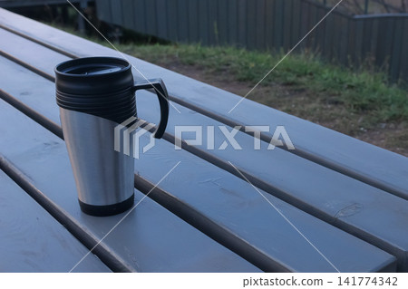 Calm Solo Moment With Stainless Steel Cup. Sunny Outdoor Scene Featuring Shiny Travel Mug By Grass. Serene Morning Outdoors With Reflective Insulated Cup Resting On Wooden Slats Near Fence Calm Solo Moment With Stainless Steel Cup. Sunny Outdoor Scene Featuring Shiny Travel Mug By Grass. Serene Morning Outdoors With Reflective Insulated Cup Resting On Wooden Slats Near Fence 141774342