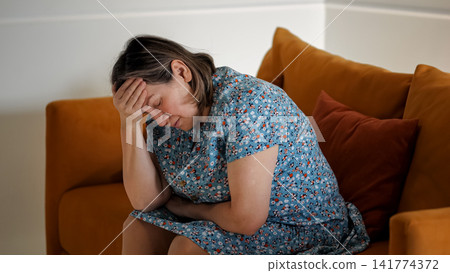 Woman Experiencing Pain While Seated Silently. Woman Shows Signs Of Stress And Headache Indoors Quietly. Woman Sitting On Sofa Exhibits Tension And Discomfort Due To Headache In Peaceful Room 141774372