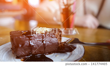 Sunlit Chocolate Cake Slice At Cafe, Lens Flare And Soft Bokeh, Glass Drink In Background, Person In Pale Sweater Blurred, Fork Resting Beside Slice, Warm Relaxed Atmosphere For Casual Gathering Sunlit Chocolate Cake Slice At Cafe, Lens Flare And Soft Bokeh, Glass Drink In Background, Person In Pale Sweater Blurred, Fork Resting Beside Slice, Warm Relaxed Atmosphere For Casual Gathering 141774405