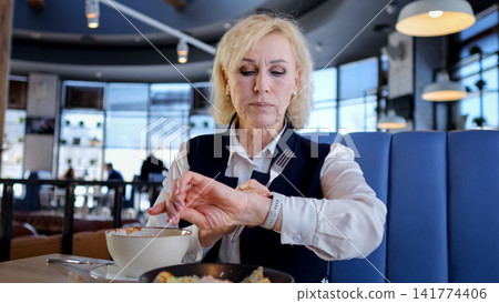 Elderly Businesswoman Checking Watch During Lunch In Modern Cafe Booth, Stirring Soup While Glancing At Wristwatch, Wearing Blazer And White Blouse, Focused Expression, Large Windows With Daylight, Elderly Businesswoman Checking Watch During Lunch In Modern Cafe Booth, Stirring Soup While Glancing At Wristwatch, Wearing Blazer And White Blouse, Focused Expression, Large Windows With Daylight, 141774406