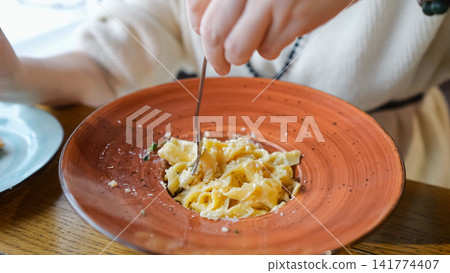 Hands Twirling Creamy Pasta At Cafe, Terracotta Bowl Topped With Parmesan And Cracked Pepper, Rich Sauce Coating Ribbons, Cozy Table Setting With Linen, Casual Sweater Sleeve Visible, Food Blogger 141774407