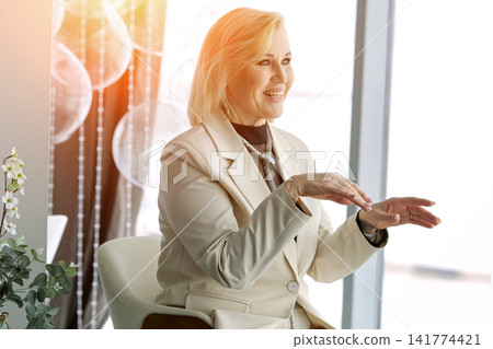 Woman Actively Gesturing During Friendly Chat. Woman Engaged In Animated Conversation Near Bright Window. Woman Energetically Gesturing And Smiling While Talking In Modern Setting Near Window Woman Actively Gesturing During Friendly Chat. Woman Engaged In Animated Conversation Near Bright Window. Woman Energetically Gesturing And Smiling While Talking In Modern Setting Near Window 141774421