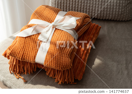 Warm Cozy Orange Textured Blanket Gift Wrapped with White Ribbon Resting on Soft Sofa Cushion Close Up Shot Warm Cozy Orange Textured Blanket Gift Wrapped with White Ribbon Resting on Soft Sofa Cushion Close Up Shot 141774637