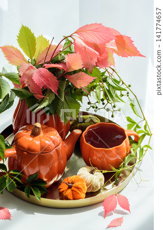 Autumn themed pumpkin teapot and cup with vibrant red and green leaves and mini pumpkins arranged on a metallic tray for seasonal decoration and cozy beverage time Autumn themed pumpkin teapot and cup with vibrant red and green leaves and mini pumpkins arranged on a metallic tray for seasonal decoration and cozy beverage time 141774657