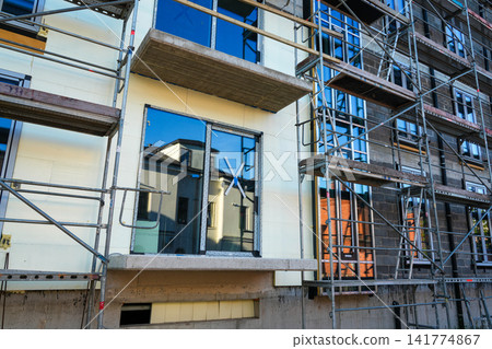Facade Insulation and Scaffolding at Modern Apartment Building Construction with New Windows Installed Facade Insulation and Scaffolding at Modern Apartment Building Construction with New Windows Installed 141774867