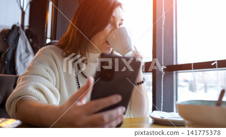Woman Sipping Coffee At Cafe While Holding Phone And Gazing Out Window, Quiet Reflective Break For Remote Worker, Warm Sunlight On Face, Steam Rising From Cup, Soft Cozy Interior And Thoughtful Mood Woman Sipping Coffee At Cafe While Holding Phone And Gazing Out Window, Quiet Reflective Break For Remote Worker, Warm Sunlight On Face, Steam Rising From Cup, Soft Cozy Interior And Thoughtful Mood 141775378