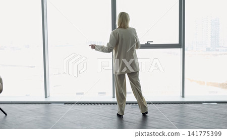 Female At Large Window Observing Skyline. Lady In Tailored Attire Examining Urban Landscape Thoughtfully. Woman In Professional Wear Gazing Over Metropolitan Horizon With Intent Focus Female At Large Window Observing Skyline. Lady In Tailored Attire Examining Urban Landscape Thoughtfully. Woman In Professional Wear Gazing Over Metropolitan Horizon With Intent Focus 141775399