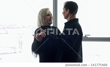 Couple Dancing By Window White Woman And White Man Share Joyful Embrace Bathed In Soft Backlight, Urban Skyline Visible Through Glass, Smiling Faces, Graceful Movement, Intimate Connection Couple Dancing By Window White Woman And White Man Share Joyful Embrace Bathed In Soft Backlight, Urban Skyline Visible Through Glass, Smiling Faces, Graceful Movement, Intimate Connection 141775400