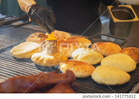 Street Food Vendor wearing black gloves Brushing oil freshly baked round Flatbreads cooking on Hot Grill. Traditional outdoor food preparation at a local festival Street Food Vendor wearing black gloves Brushing oil freshly baked round Flatbreads cooking on Hot Grill. Traditional outdoor food preparation at a local festival 141775812
