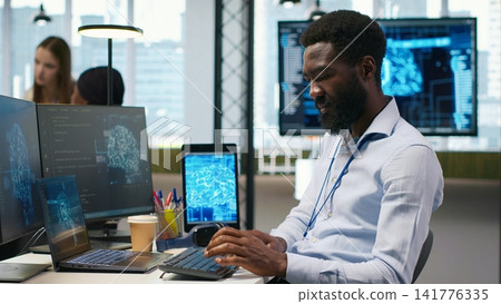 System administrator at office desk developing software using AI, listening music. IT expert wearing audio headset working on computer, debugging applications with artificial intelligence, camera A System administrator at office desk developing software using AI, listening music. IT expert wearing audio headset working on computer, debugging applications with artificial intelligence, camera A 141776335