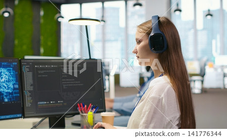 Junior dev developing software applications, optimizing code for performance, listening music. Young female engineer wearing headphones in office using AI to resolve bottlenecks, camera A Junior dev developing software applications, optimizing code for performance, listening music. Young female engineer wearing headphones in office using AI to resolve bottlenecks, camera A 141776344