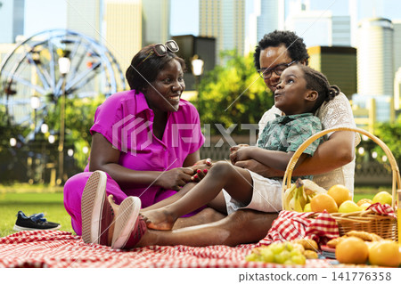 Happy parents engage in playful conversation with expressive child while relaxing together beside fruits and picnic food on checkered blanket. Family leisure on sunny day in urban park. Happy parents engage in playful conversation with expressive child while relaxing together beside fruits and picnic food on checkered blanket. Family leisure on sunny day in urban park. 141776358