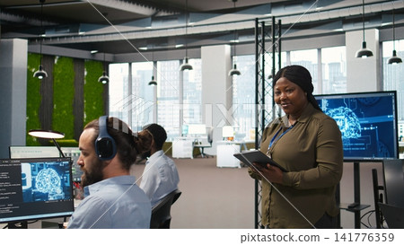 Smiling female engineer next to office coworkers developing software applications, integrating AI automation. Happy woman next to developers at work debugging applications, camera A Smiling female engineer next to office coworkers developing software applications, integrating AI automation. Happy woman next to developers at work debugging applications, camera A 141776359